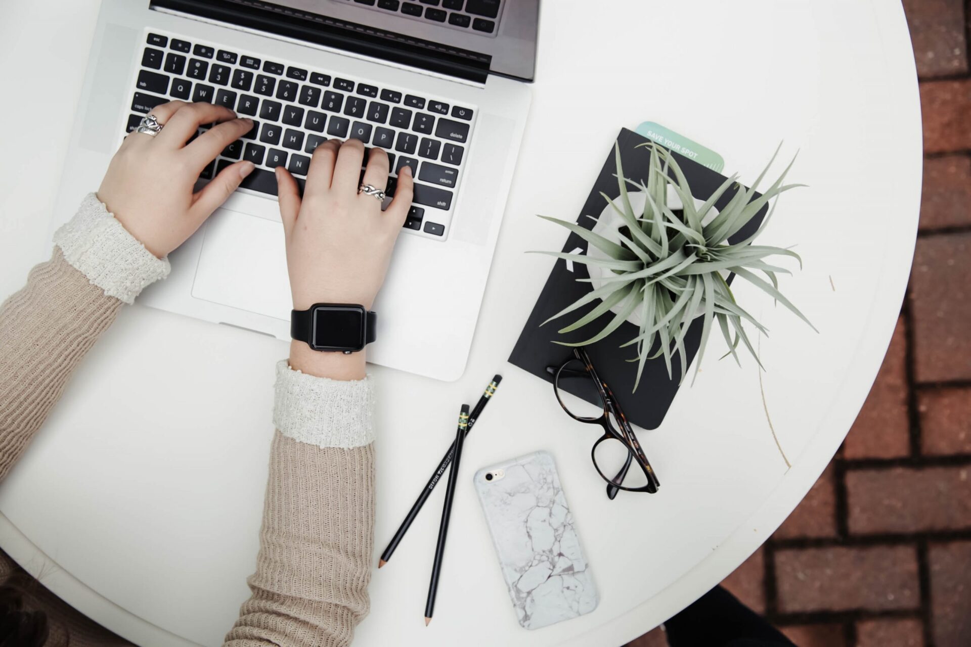 A top-down view of a white circular table with a person working on a laptop. The person is wearing a beige sweater and a smartwatch. On the table, next to the laptop, are a smartphone with a marble case, a pair of glasses, two pencils, and a small potted plant.