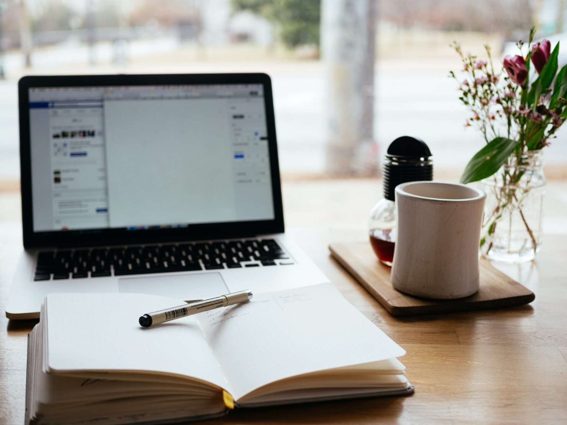 A cozy workspace with a laptop, an open notebook, and a pen on a wooden table. Next to the notebook is a white coffee mug on a wooden coaster, a small glass vase with fresh flowers, and a French press coffee maker.