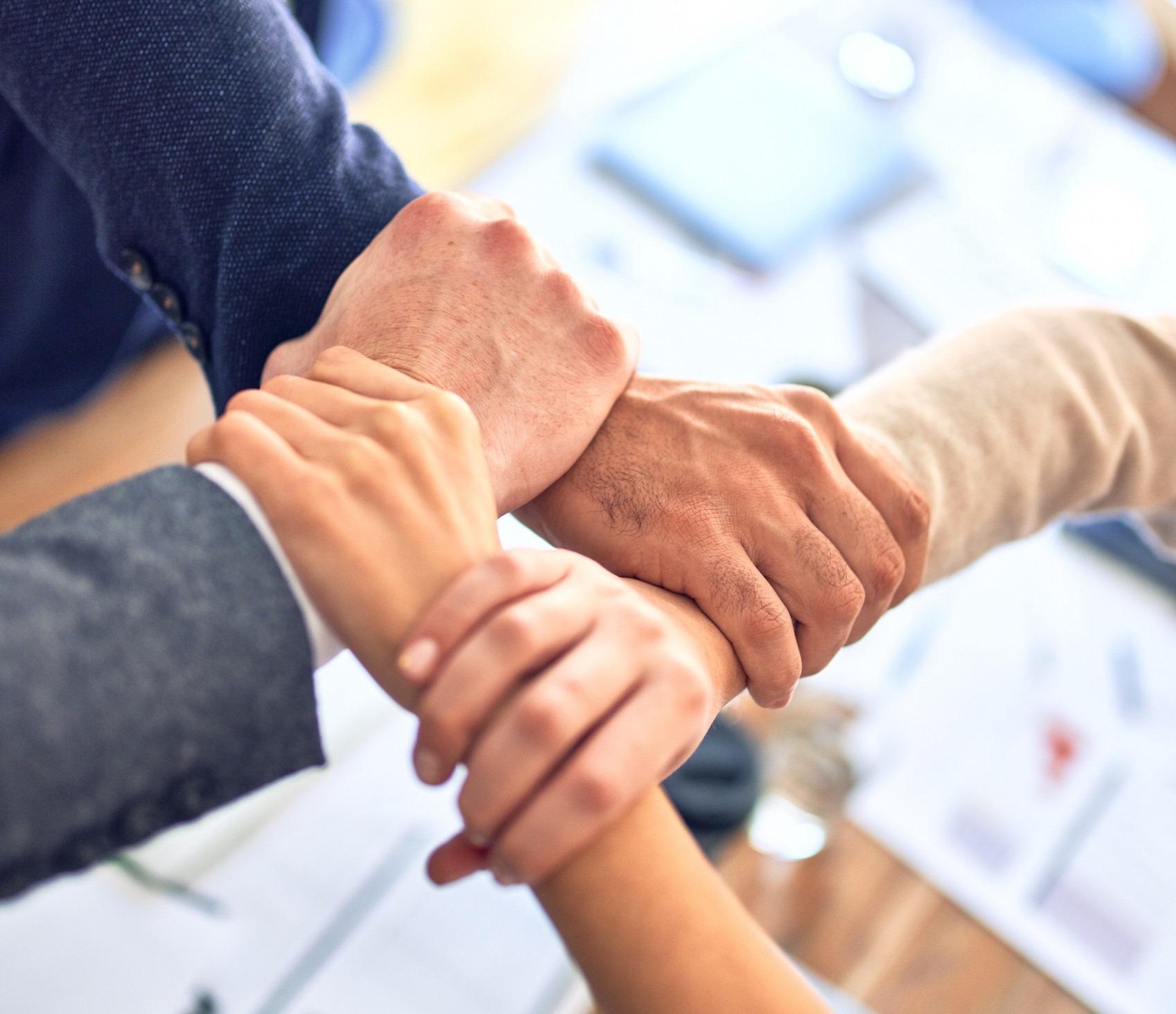 Four diverse hands are joined together in a central knot, symbolizing unity and teamwork.