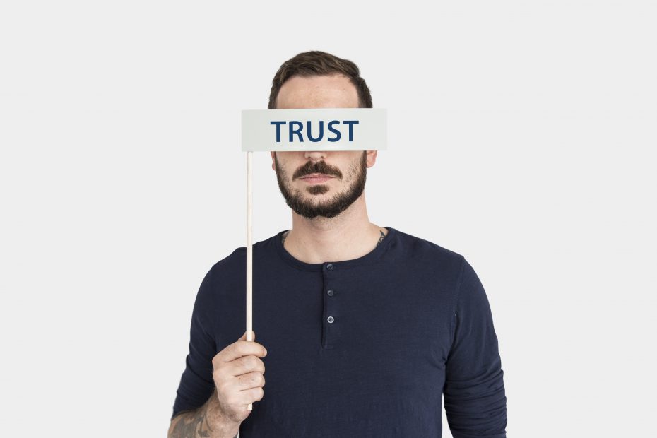 A man with a beard and wearing a dark blue shirt holds up a sign in front of his eyes that reads "TRUST." The background is plain white, making the text on the sign and the man's expression the focal points of the image. The overall composition conveys a message about trust and perception. A man with a beard and wearing a dark blue shirt holds up a sign in front of his eyes that reads "TRUST." The background is plain white, making the text on the sign and the man's expression the focal points of the image. The overall composition conveys a message about trust and perception.