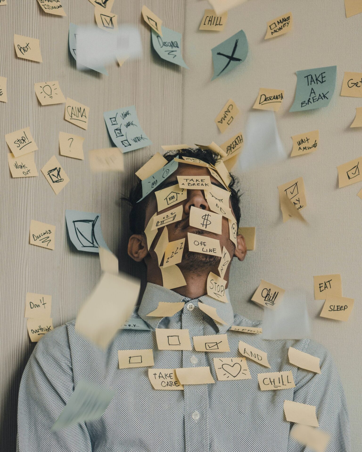 A photograph of a person sitting against a wall, covered in numerous sticky notes. The sticky notes are scattered all over the person's face, body, and the wall around them, each containing various reminders and tasks. The person appears overwhelmed and stressed, with eyes closed and head slightly tilted back. The background is neutral, emphasizing the chaotic scene created by the sticky notes. The overall mood reflects a sense of being overwhelmed by tasks and responsibilities.
