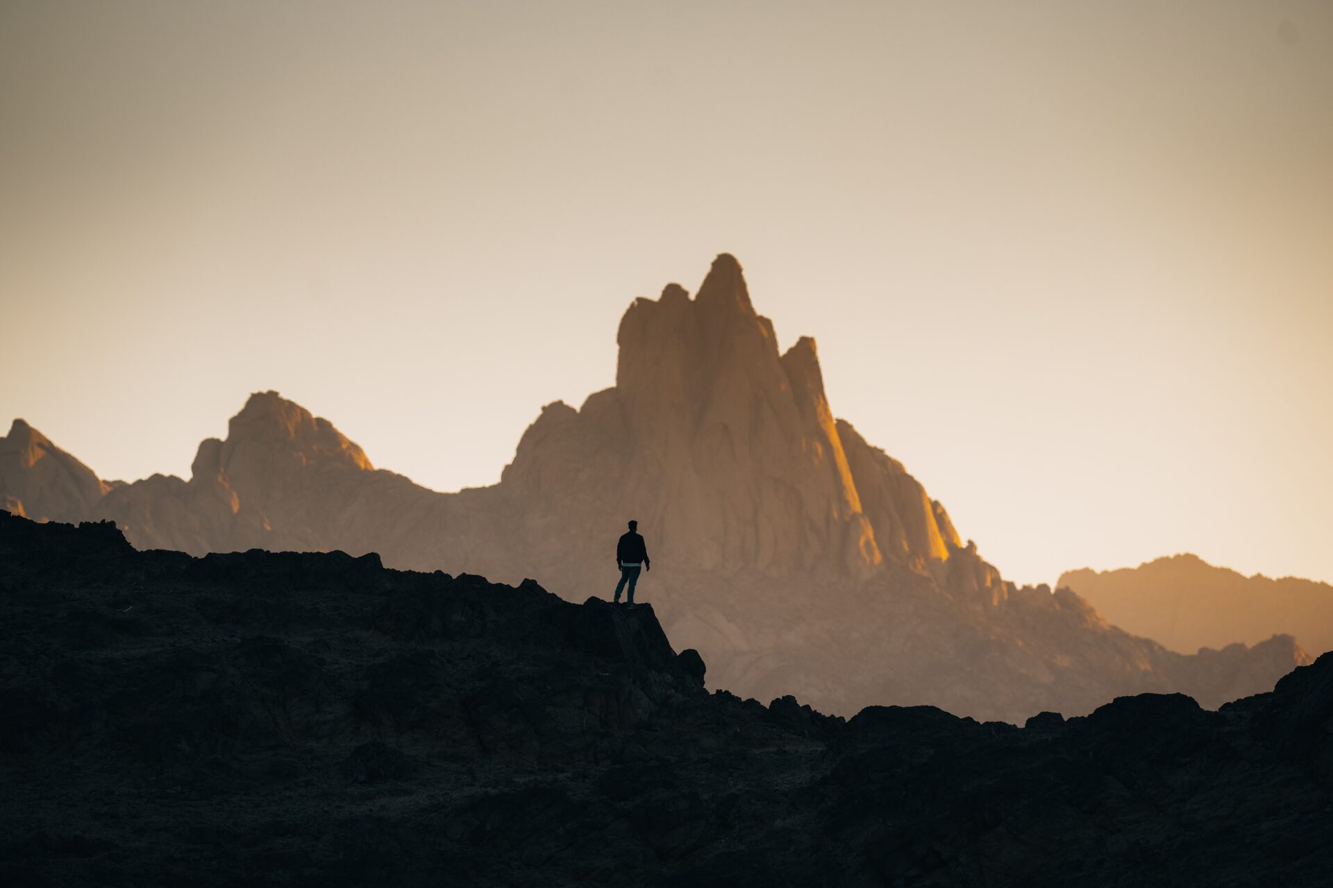 A photograph of a lone hiker standing on a rocky terrain with a dramatic mountain range in the background. The sun is setting or rising, casting a golden glow on the jagged peaks and creating a silhouette effect on the hiker and the foreground. The scene exudes a sense of adventure, solitude, and the grandeur of nature, with the warm light enhancing the rugged beauty of the landscape.