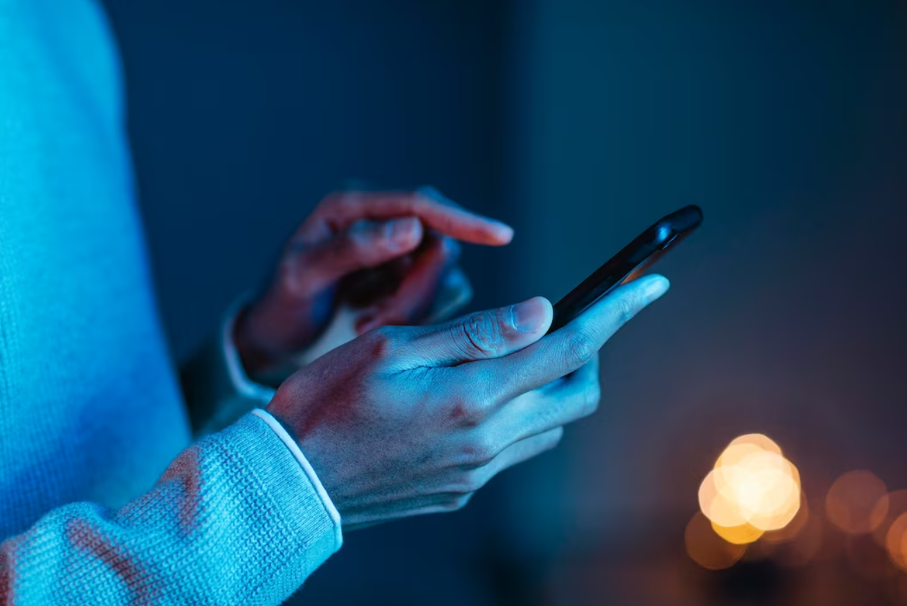 The image shows a close-up of a person using a smartphone, with both hands interacting with the device. The person is wearing a light-colored sweater, and the background is dimly lit with a soft, warm light in the distance, creating a cozy and intimate atmosphere. The focus is on the hands and the smartphone, emphasizing the act of texting, browsing, or engaging with digital content.