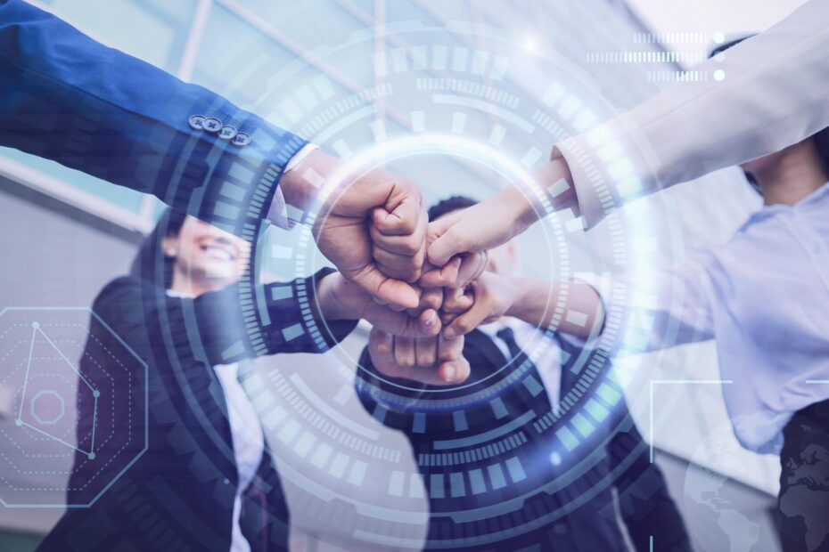 A photograph of a group of people standing in a circle, each extending a fist towards the center to create a collective fist bump. They are dressed in business attire, indicating a professional setting. Overlaid on the image are translucent, futuristic digital graphics, including circular HUD (Heads-Up Display) elements and various data icons, suggesting themes of teamwork, technology, and innovation. The background shows a modern building with glass windows, adding to the contemporary and dynamic feel of the scene.