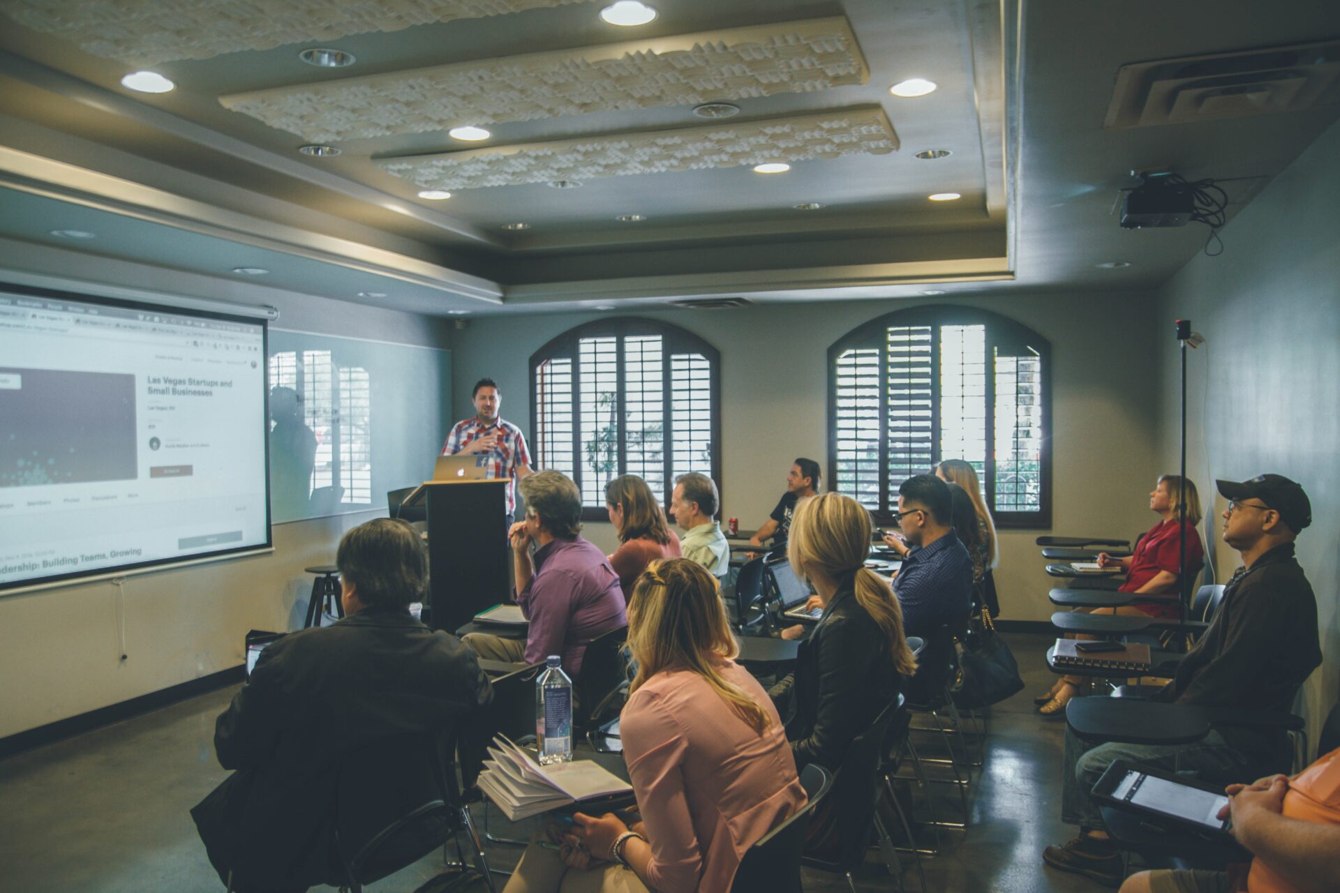 A photograph of a classroom or seminar room filled with people seated and listening attentively to a speaker at the front. The speaker is standing next to a podium and a large screen displaying a presentation. The room has a modern design with arched windows, letting in natural light, and a high ceiling with recessed lighting. The audience appears engaged, some taking notes or using laptops. The overall atmosphere is professional and focused, indicating a learning or training session.