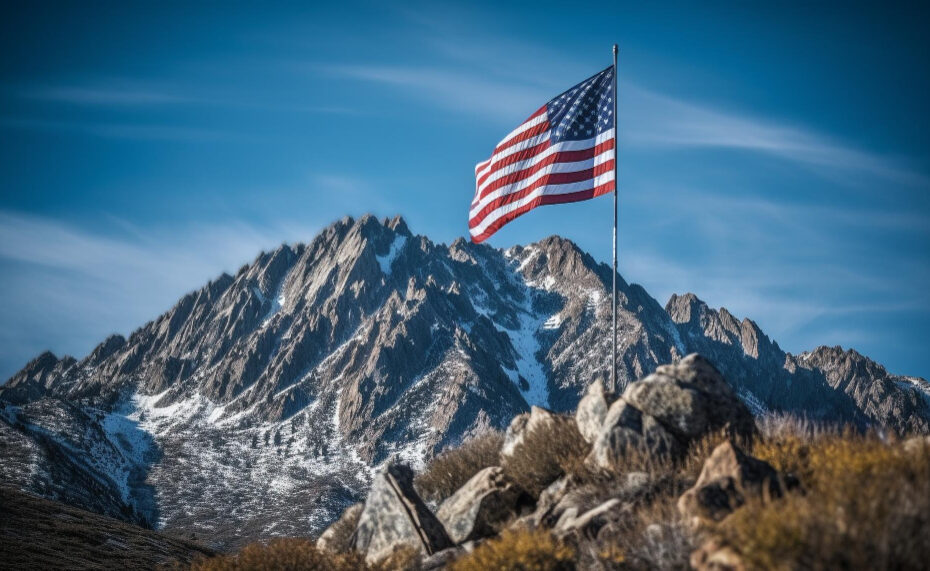 An image of the USA flag raised on a flagpole at the summit of a mountain, with a clear blue sky in the background
