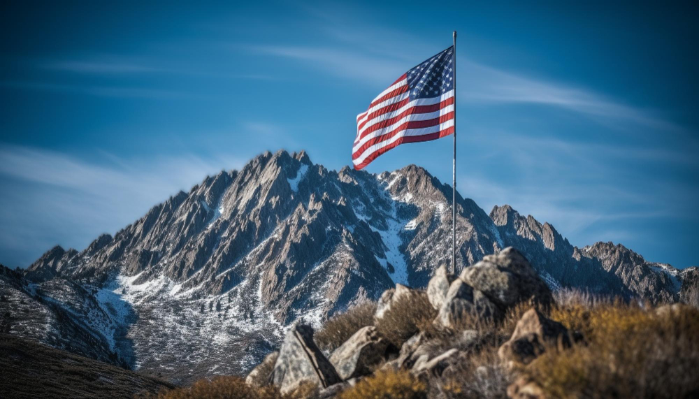 An image of the USA flag raised on a flagpole at the summit of a mountain, with a clear blue sky in the background