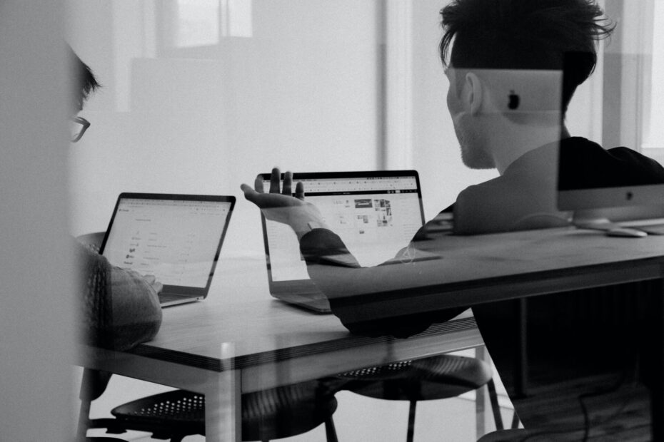 Two employees sitting at the same table with two laptops in front of them and having a discussion.