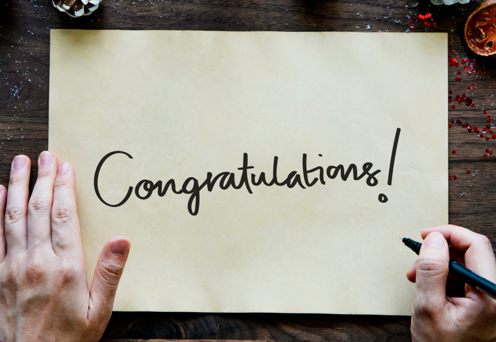 An image of a human hand holding a pen in front of a large piece of paper with the word "Congratulations!" written on it.