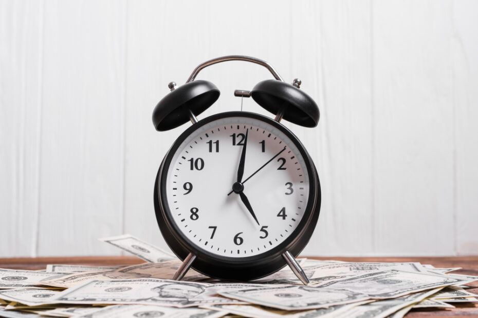 Image of a small analog clock centered with stacks of money placed underneath, symbolizing the connection between time and finances.