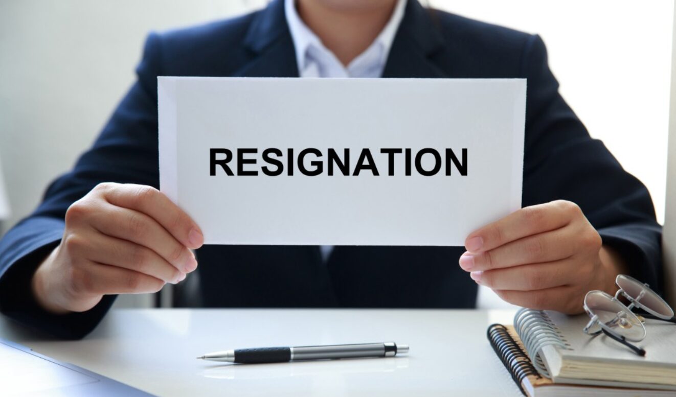 The image shows a person in a business suit holding a sign that reads "RESIGNATION." The individual is seated at a desk with a pen, notebook, and glasses, indicating a formal setting. The image clearly conveys the act of resigning from a job, emphasizing a significant and serious professional decision.