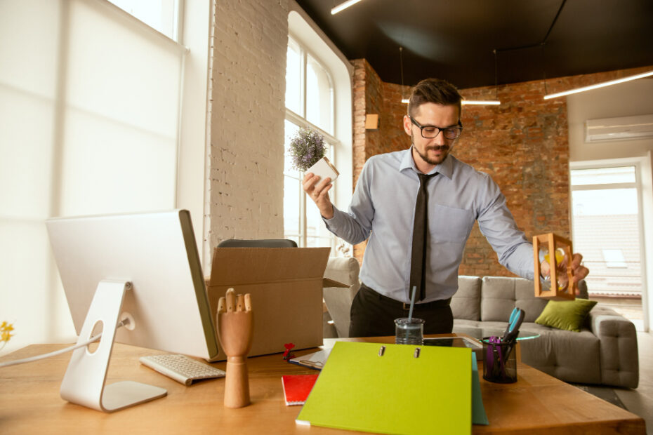 The image depicts a man in a modern office setting, organizing or tidying up his desk. He is holding a small potted plant in one hand and a picture frame in the other. The desk features a computer, a stand with a hand model, colorful folders, and various office supplies. The office environment is stylish, with large windows allowing natural light to fill the space, exposed brick walls, and contemporary furniture, creating a productive and aesthetically pleasing atmosphere.