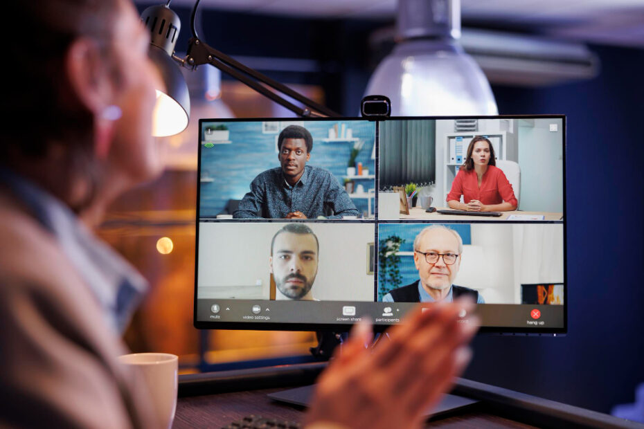 A person participating in a virtual meeting, visible from behind and clapping their hands. The monitor displays a video conference with four colleagues in different locations, each in their own video frame. The room is dimly lit with a desk lamp illuminating the workspace, creating a cozy and focused atmosphere.