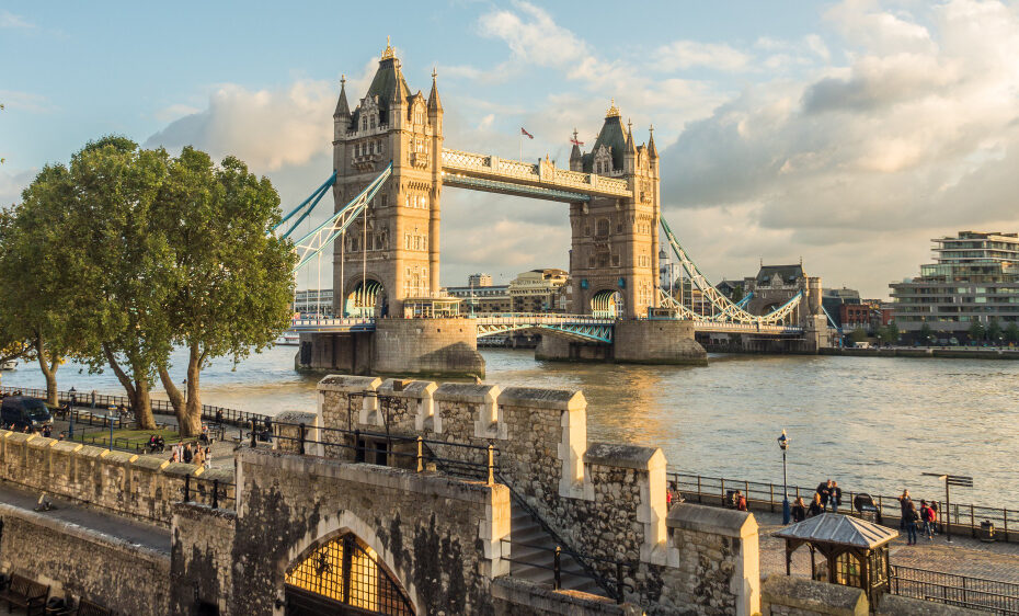 The image shows Tower Bridge in London, a historic suspension bridge over the River Thames. The scene is set during the day with a partly cloudy sky. The bridge's two towers are prominent, and the bridge itself is slightly raised. In the foreground, there are trees, stone walls, and a riverside walkway where people are strolling and relaxing. The river is calm, reflecting the warm, golden sunlight. On the far side of the river, modern buildings and greenery are visible.