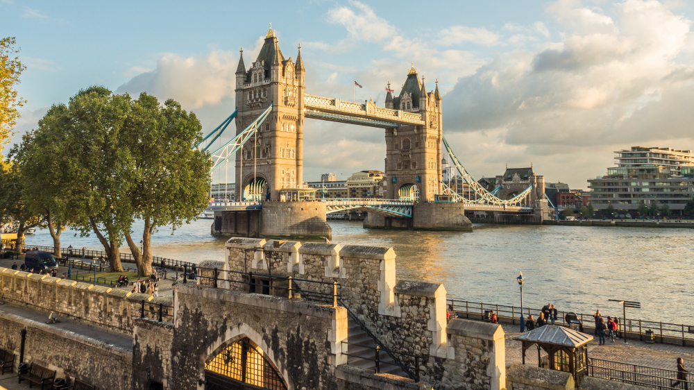 The image shows Tower Bridge in London, a historic suspension bridge over the River Thames. The scene is set during the day with a partly cloudy sky. The bridge's two towers are prominent, and the bridge itself is slightly raised. In the foreground, there are trees, stone walls, and a riverside walkway where people are strolling and relaxing. The river is calm, reflecting the warm, golden sunlight. On the far side of the river, modern buildings and greenery are visible.