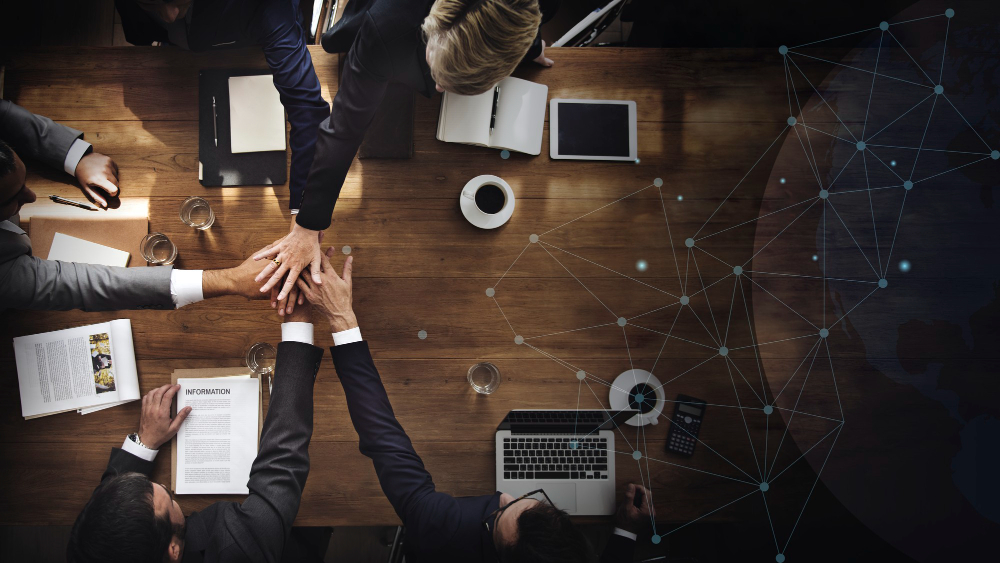 A group of business professionals is seated around a large wooden table, engaging in a collaborative meeting. The individuals are placing their hands together in a gesture of unity and teamwork, signifying collaboration and shared goals. The table is cluttered with open laptops, documents, notebooks, and cups of coffee, indicating a productive work session. In the background, a digital network overlay with connected dots and lines symbolizes global communication and interconnectedness in the business world.