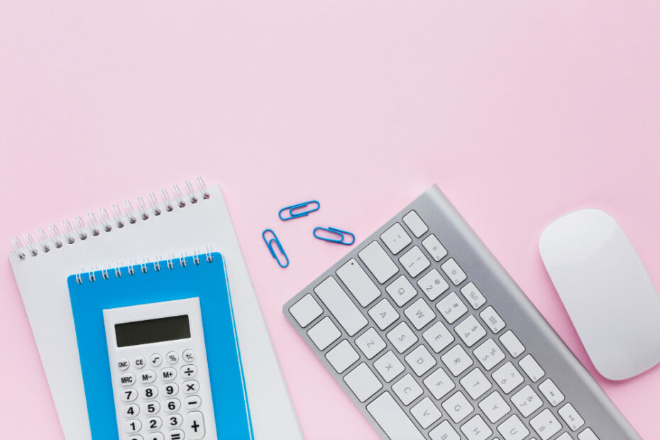 A neatly arranged workspace on a pastel pink background featuring a white wireless keyboard, a white wireless mouse, two spiral notebooks, and a blue calculator placed on top of the notebooks. A few blue paper clips are scattered nearby, adding a touch of color coordination. The setup is clean and organized, evoking a sense of simplicity and productivity.