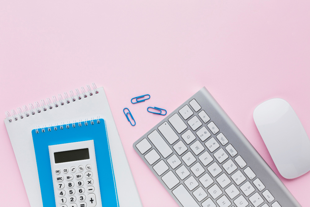 A neatly arranged workspace on a pastel pink background featuring a white wireless keyboard, a white wireless mouse, two spiral notebooks, and a blue calculator placed on top of the notebooks. A few blue paper clips are scattered nearby, adding a touch of color coordination. The setup is clean and organized, evoking a sense of simplicity and productivity.