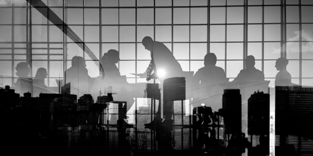 A black and white image of a group of people in a modern office, with large windows overlooking a city skyline. The scene is captured with reflections of the city superimposed on the silhouettes of the individuals, creating a layered, abstract effect. The group appears to be engaged in a meeting or discussion, with one person standing and addressing the others as sunlight streams in from the background.