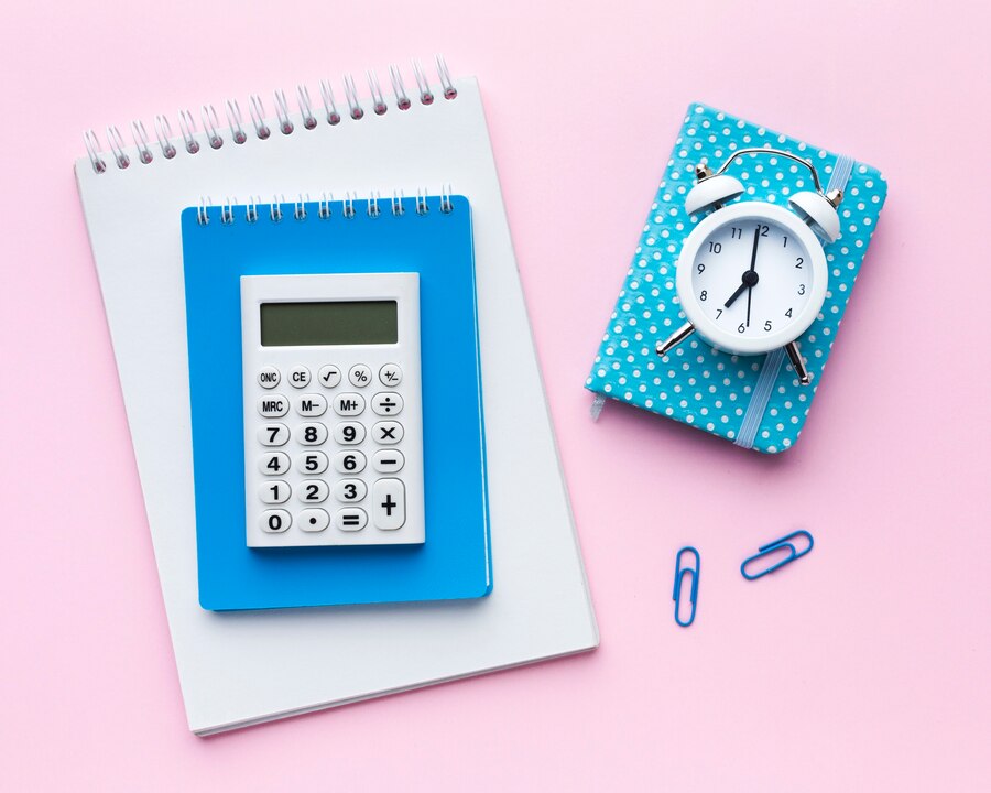 This image features a flat lay arrangement of stationery items on a pastel pink background. At the center is a white calculator resting on a blue notebook, placed on top of a white spiral-bound notepad. To the right, a small blue polka-dotted planner lies with a white alarm clock sitting on top. Blue paper clips add subtle details, creating a clean, organized, and aesthetically pleasing workspace vibe.