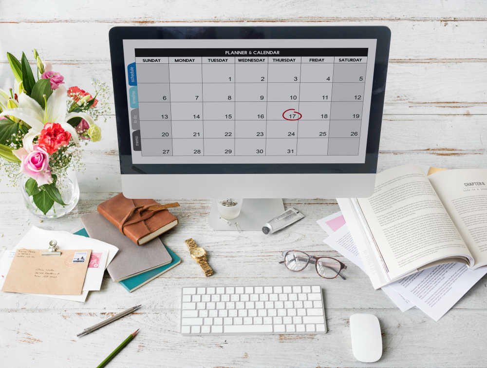A modern workspace featuring a desktop computer displaying a digital planner and calendar, with a date circled. The desk is decorated with flowers, books, glasses, stationery, and office supplies. The setup suggests the use of a vacation tracking tool for managing schedules and planning time off efficiently.