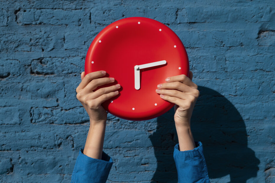 Person holding a red clock showing 3:00 against a blue brick wall, symbolizing the concept of military time and the 24-hour clock system.